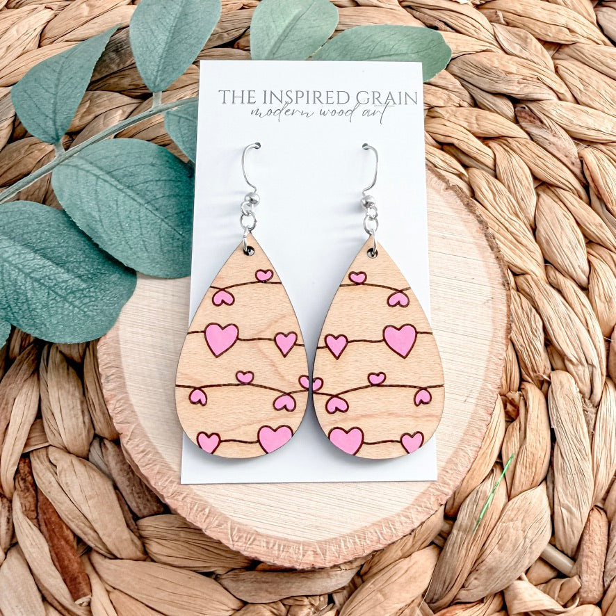 Wooden teardrop earrings with pink heart patterns on a 'The Inspired Grain' branded card, placed on a woven surface with leaves.