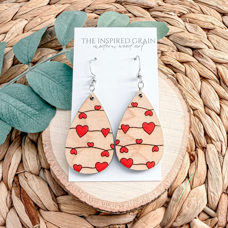 Wooden teardrop earrings with red heart patterns on a 'The Inspired Grain' card, placed on a woven surface with leaves.