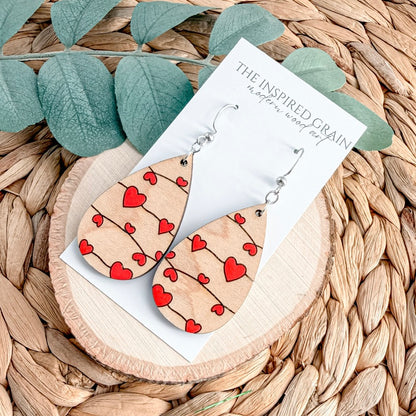 Wooden earrings with heart patterns on a branded card, placed on a woven surface with leaves.