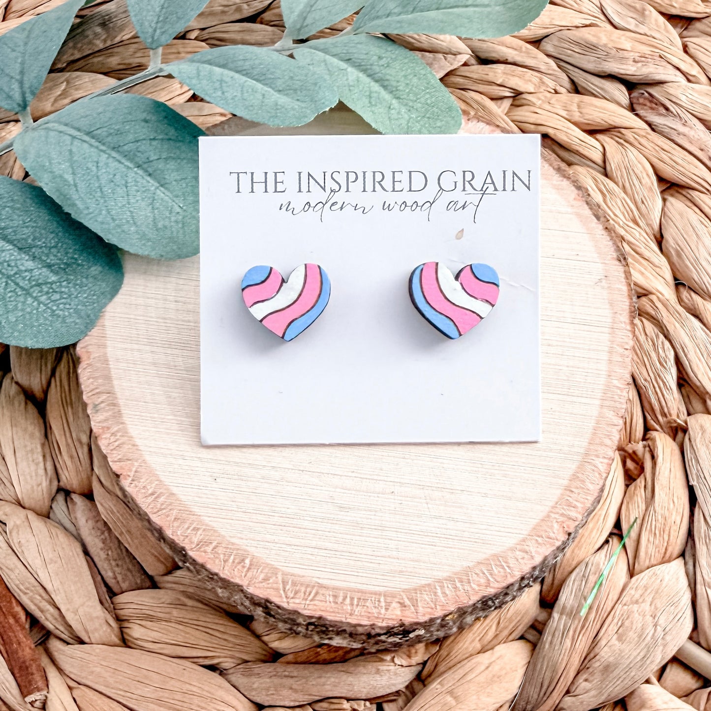 Heart-shaped earrings on a card with 'The Inspired Grain' branding, placed on a woven surface with leaves.